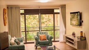 A living room with a sofa, armchair, and coffee table, a TV mounted on the wall, and a balcony with greenery visible through the windows. - Sand Grains Residences 002 (Entebbe)