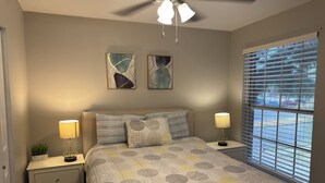 A bedroom with a bed, two bedside tables, a ceiling fan, and a window with blinds. - I042 – Upgraded 2BD Apt | Pool | DFW South 183/161 (Irving)