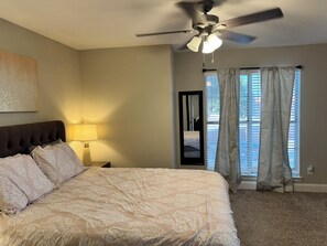 Une chambre à coucher comprenant un lit, un ventilateur de plafond, une table de chevet avec une lampe, une fenêtre avec des rideaux et une porte.