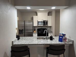 A compact kitchen with stainless steel appliances, a granite countertop, and two bar stools.