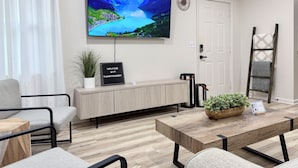 Una sala de estar moderna con un televisor de pantalla plana, un cartel de bienvenida, una planta en maceta, una mesa de centro de madera y una escalera apoyada contra la pared.