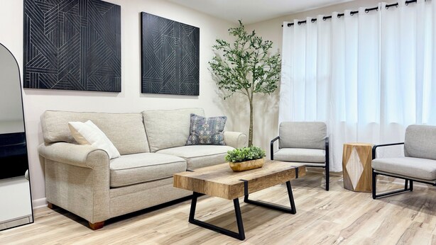 Una sala moderna con un sofá beige, una mesa de centro de madera y dos sillones. Hay dos cuadros abstractos en la pared y una planta en maceta.