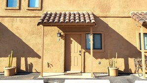 Un edificio con fachada de estuco color beige, techo de tejas y una puerta con el número '3'. A la derecha, hay dos cactus en macetas y un banco.
