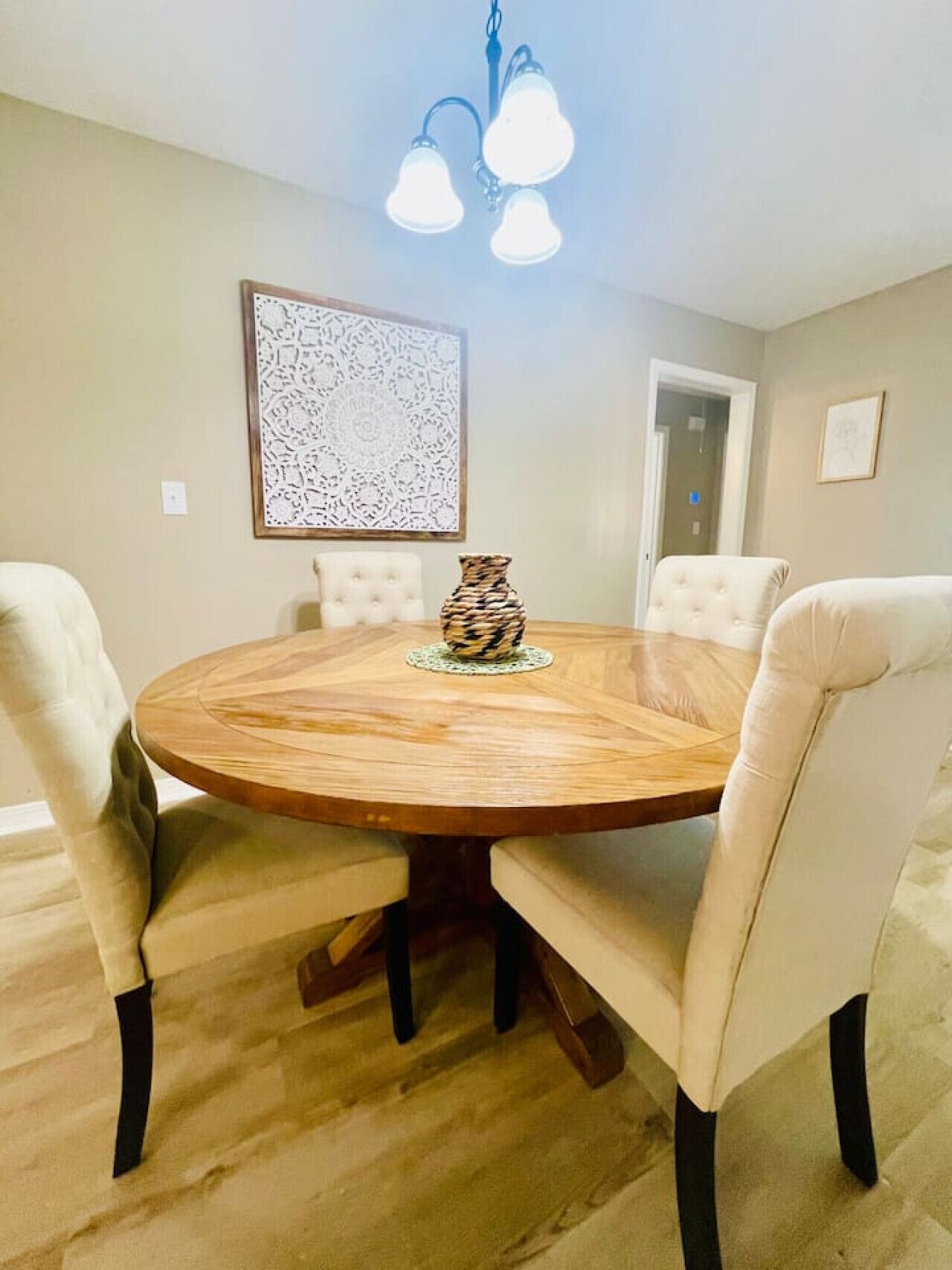 Una mesa de comedor redonda de madera con cuatro sillas tapizadas, un jarrón decorativo y una araña.