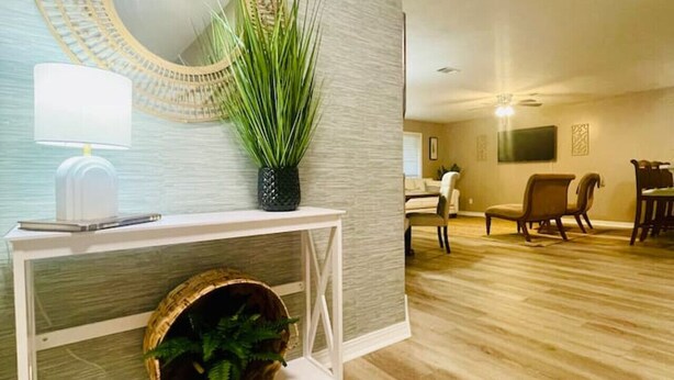 A modern living room with a light wood floor, a white console table with a plant, a round mirror, and a lamp. There is a seating area with a sofa and chairs in the background. - EastGate 4A Cozy family retreat close to downtown Hammond (Hammond)
