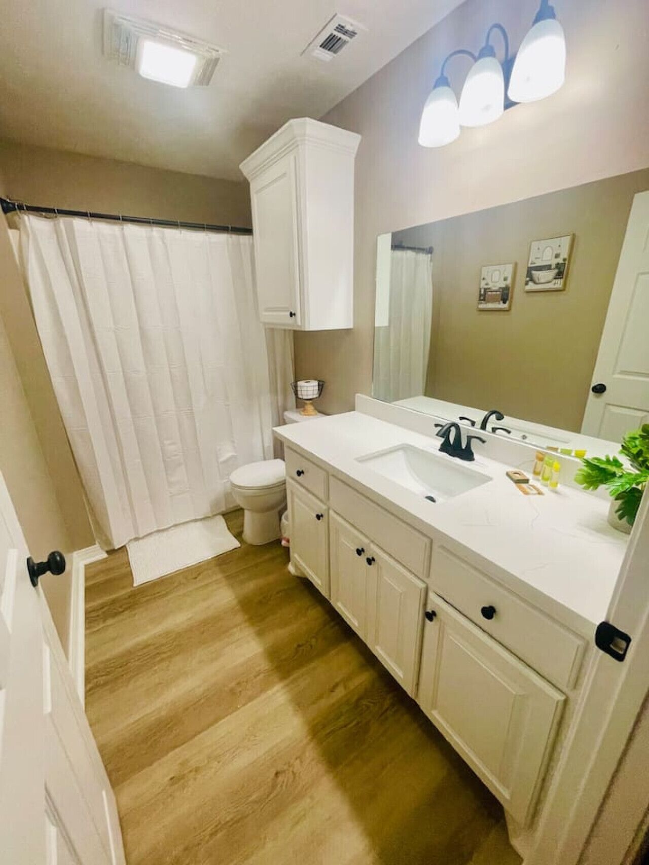 A bathroom with a wooden floor, white fixtures, a shower curtain, a toilet, and a sink with a mirror.