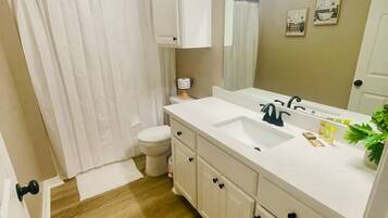 A bathroom with a wooden floor, white fixtures, a shower curtain, a toilet, and a sink with a mirror.