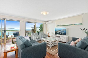 Ein modernes Wohnzimmer mit großem Fernseher, grauen Sofas, einem Couchtisch und einem Balkon mit Meerblick.