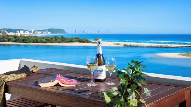 A wooden table with a bottle of champagne, two glasses, and a book on a balcony overlooking a beach and city skyline.