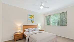 A bedroom with a bed, nightstands, a ceiling fan, a window with blinds, and a painting of a palm tree.