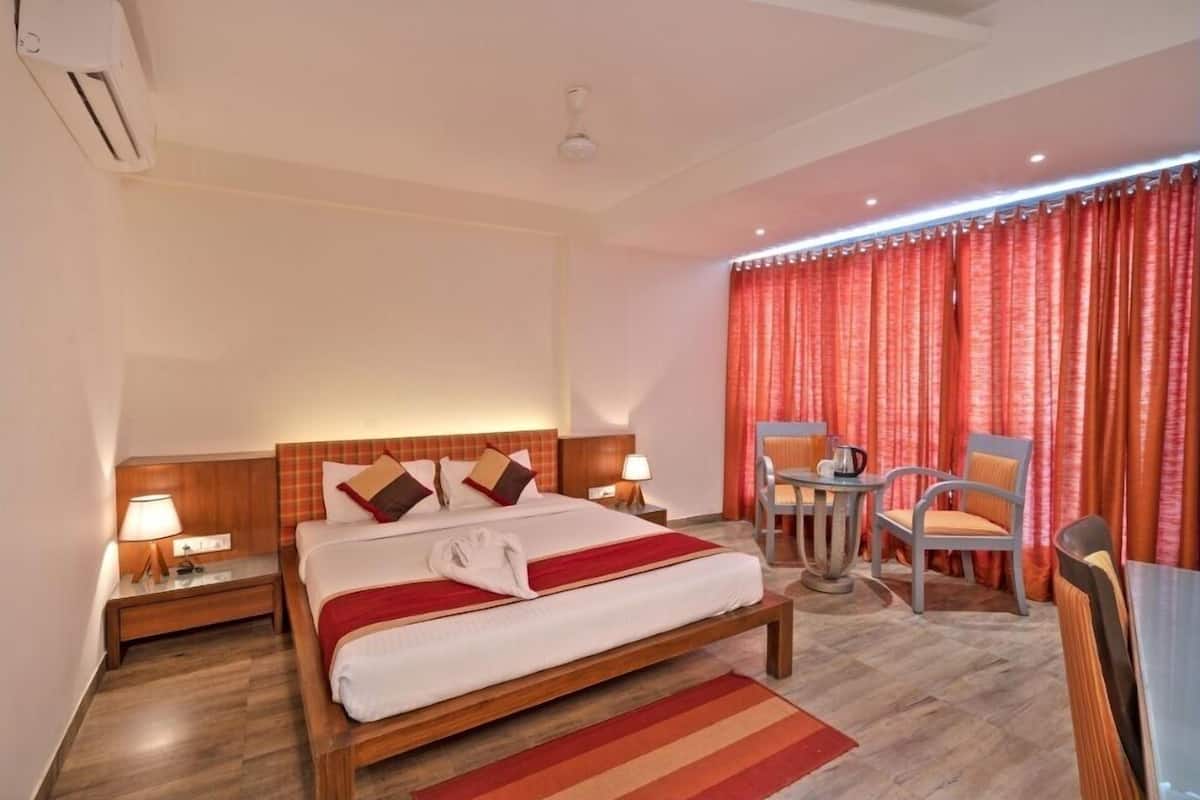 A hotel room with a large bed, a bedside table with a lamp, a small round table with chairs, and a window with red curtains.