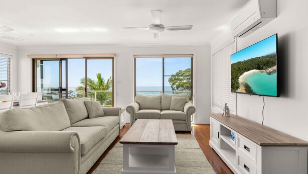 A modern living room with a sofa, a coffee table, and a TV. There are large windows with a view of the sea.