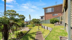 A residential backyard with a brick house, a green fence, and various plants.