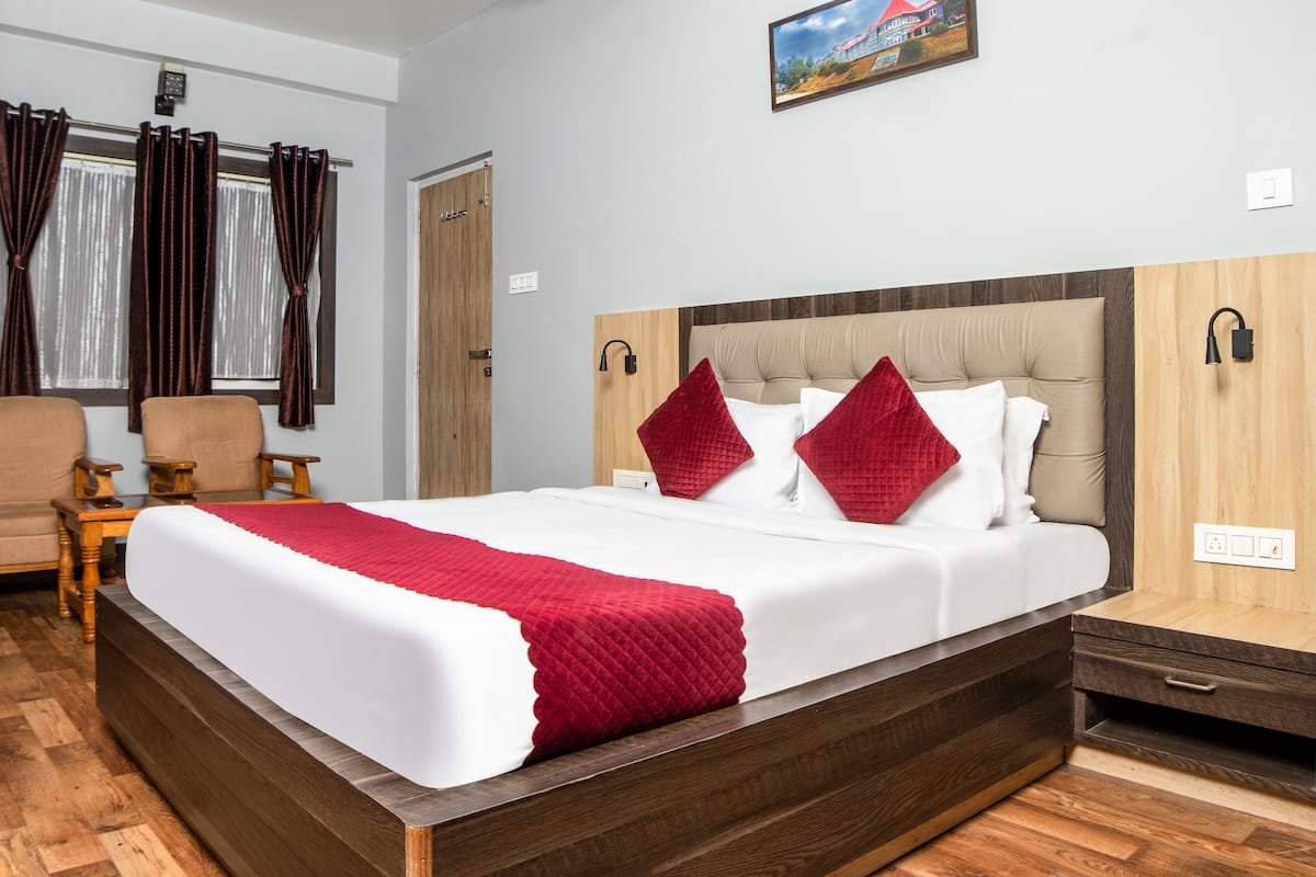 A hotel room with a large bed, wooden headboard, two armchairs, a wooden nightstand, and a painting on the wall.