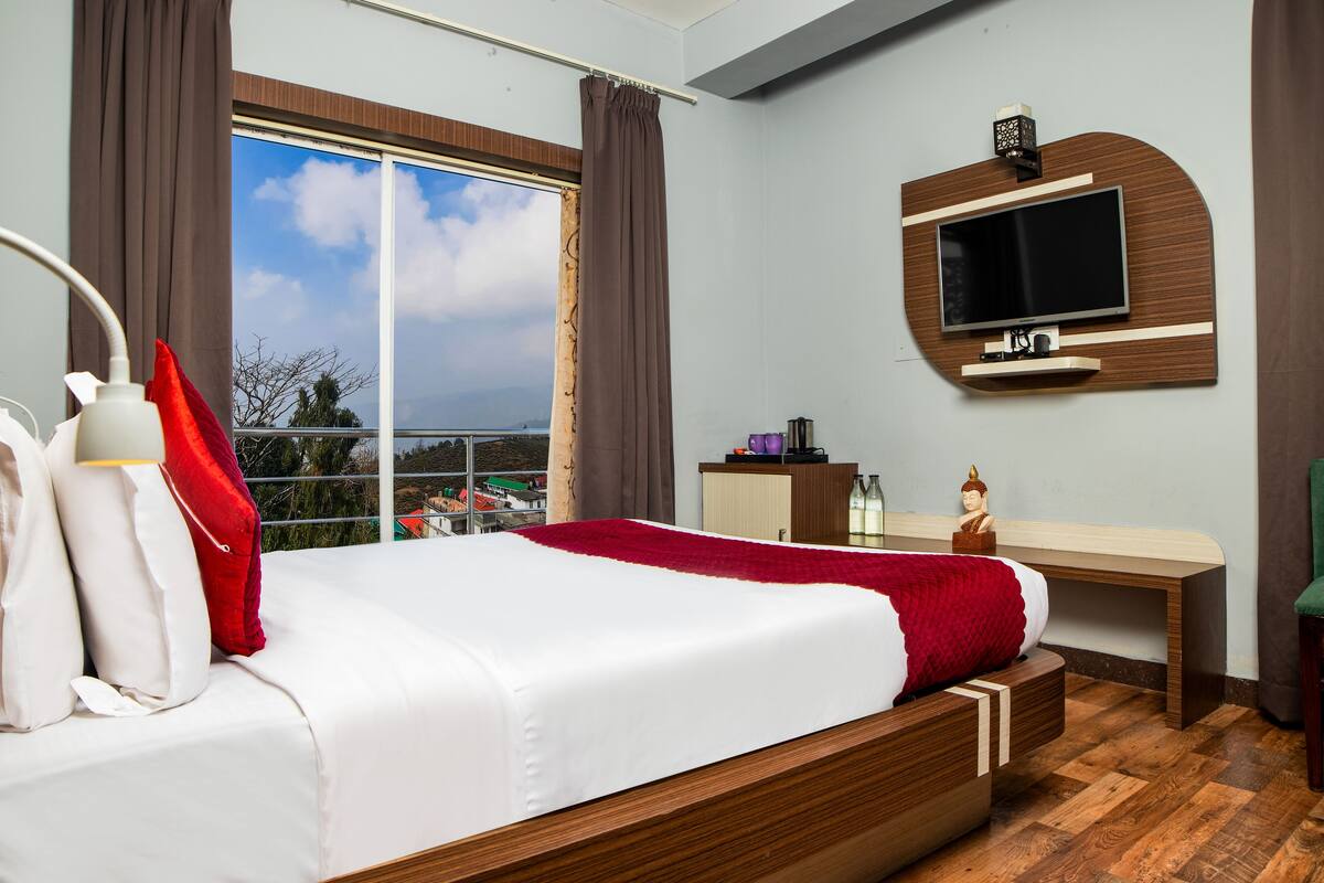 A hotel room with a large bed, a TV mounted on the wall, a desk, and a view of the city through the window.