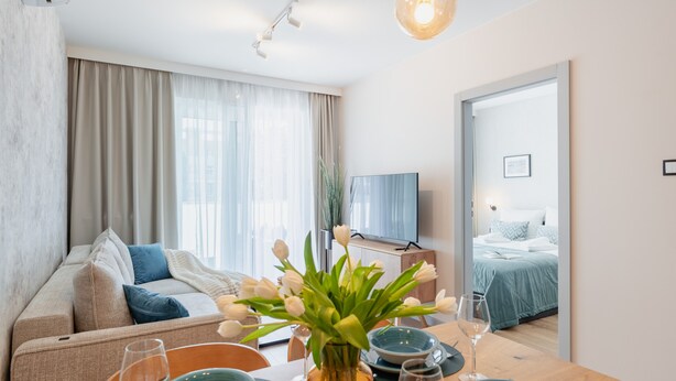 A modern hotel room with a sofa, a dining table with a vase of flowers, a TV, and a bed with a blue bedspread. - Apartamenty Sun & Snow Perłowa Przystań Kołobrzeg (Kolobrzeg)