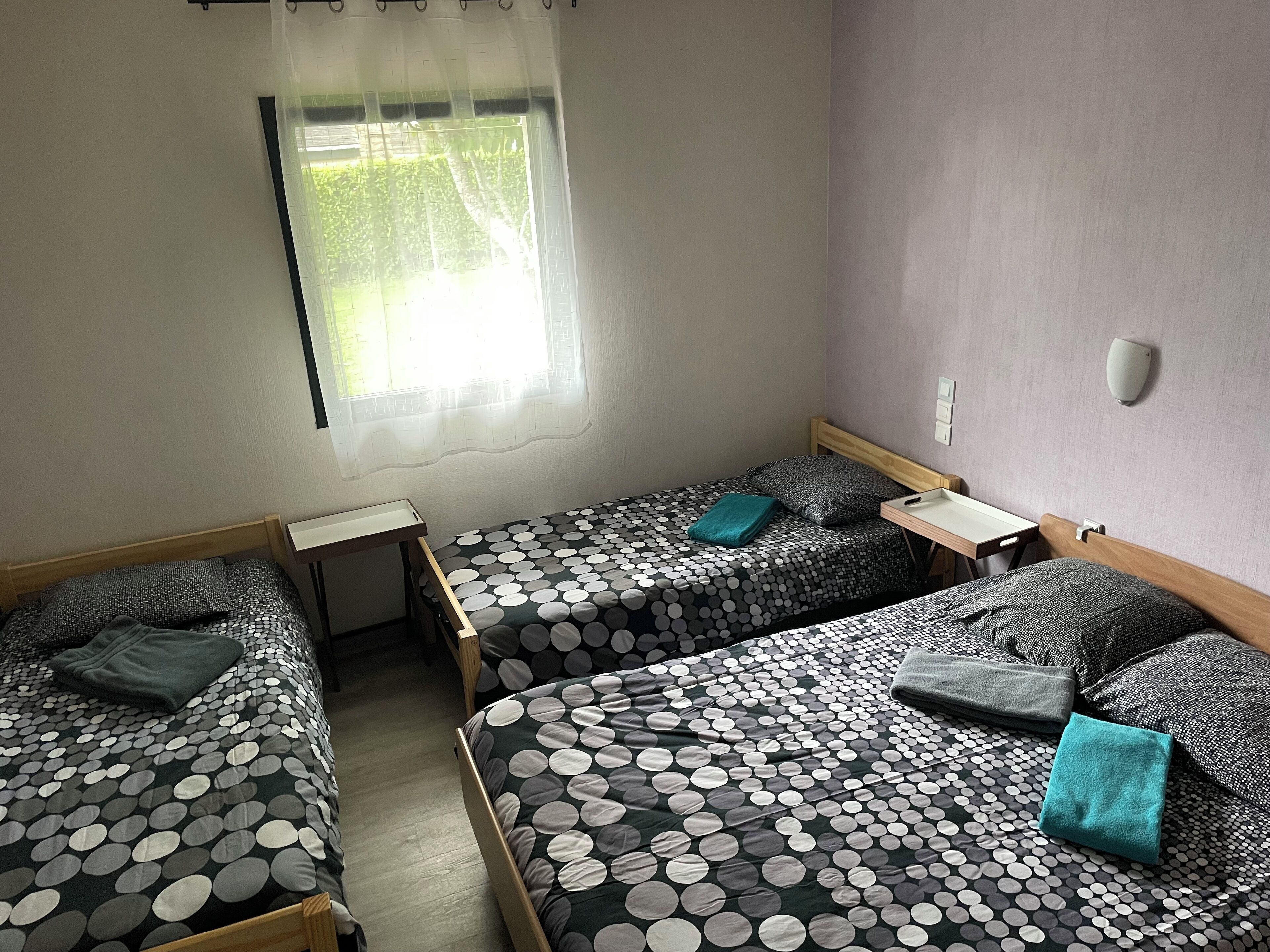 Une chambre avec deux lits simples, chacun recouvert d’une couvre-lit à motifs et agrémenté d’un oreiller. Une petite table se trouve entre les lits, et il y a une fenêtre avec un rideau.