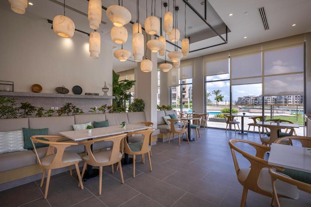 A modern indoor dining area with wooden tables and chairs, pendant lighting, and large windows offering a view of the outdoors.