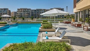 A swimming pool with lounge chairs and umbrellas, surrounded by greenery and modern buildings in the background.