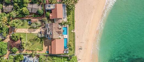 An aerial view of a coastal property with a swimming pool, beach access, and lush greenery.
