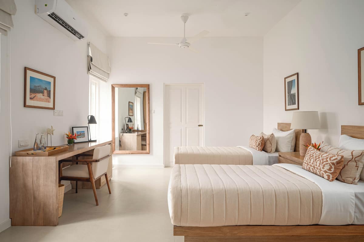 A hotel room with a wooden desk, two beds with beige bedding, a ceiling fan, and a large mirror.