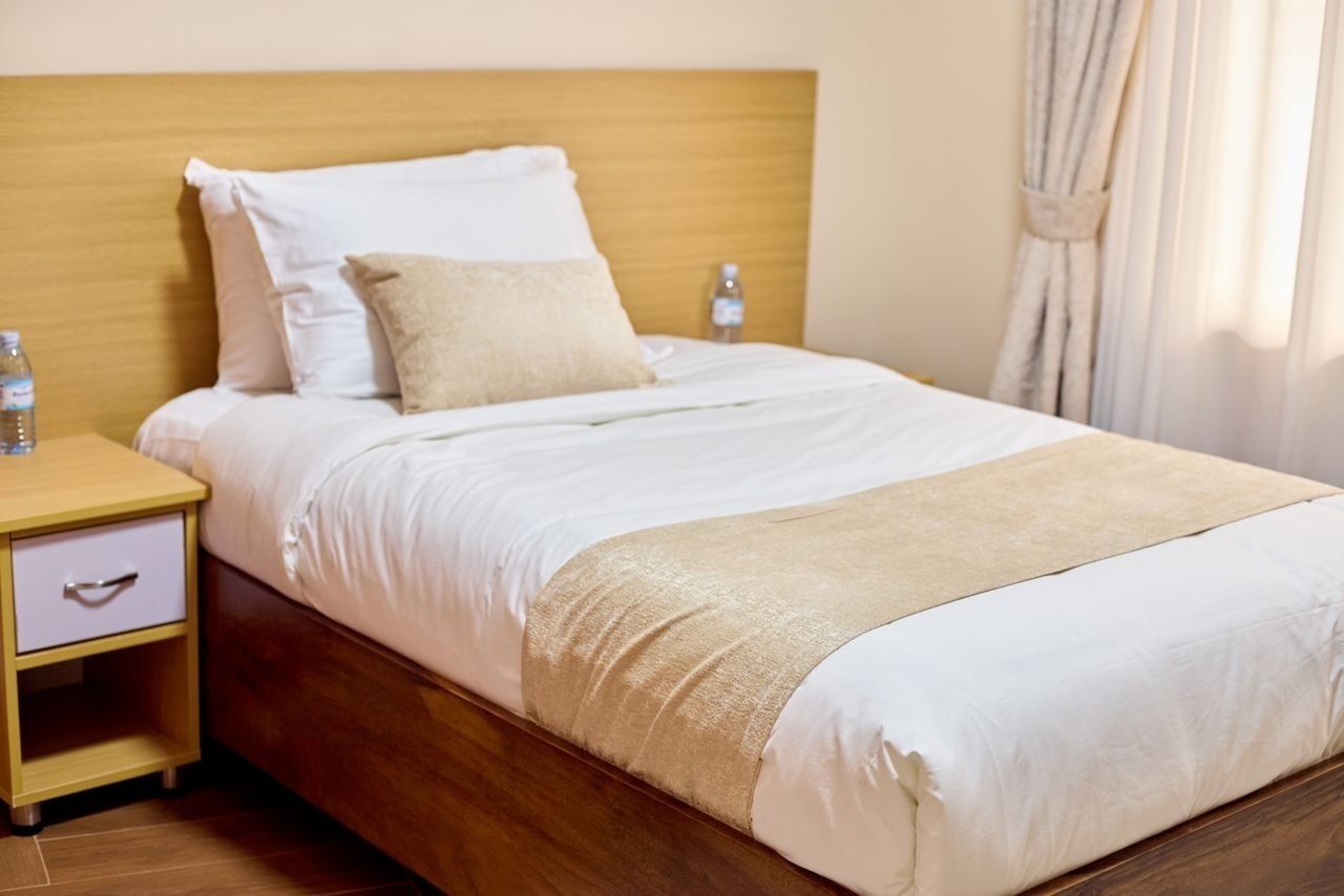 A neatly made bed with white and beige bedding, a wooden headboard, a nightstand with a drawer, and a bottle of water on the nightstand.