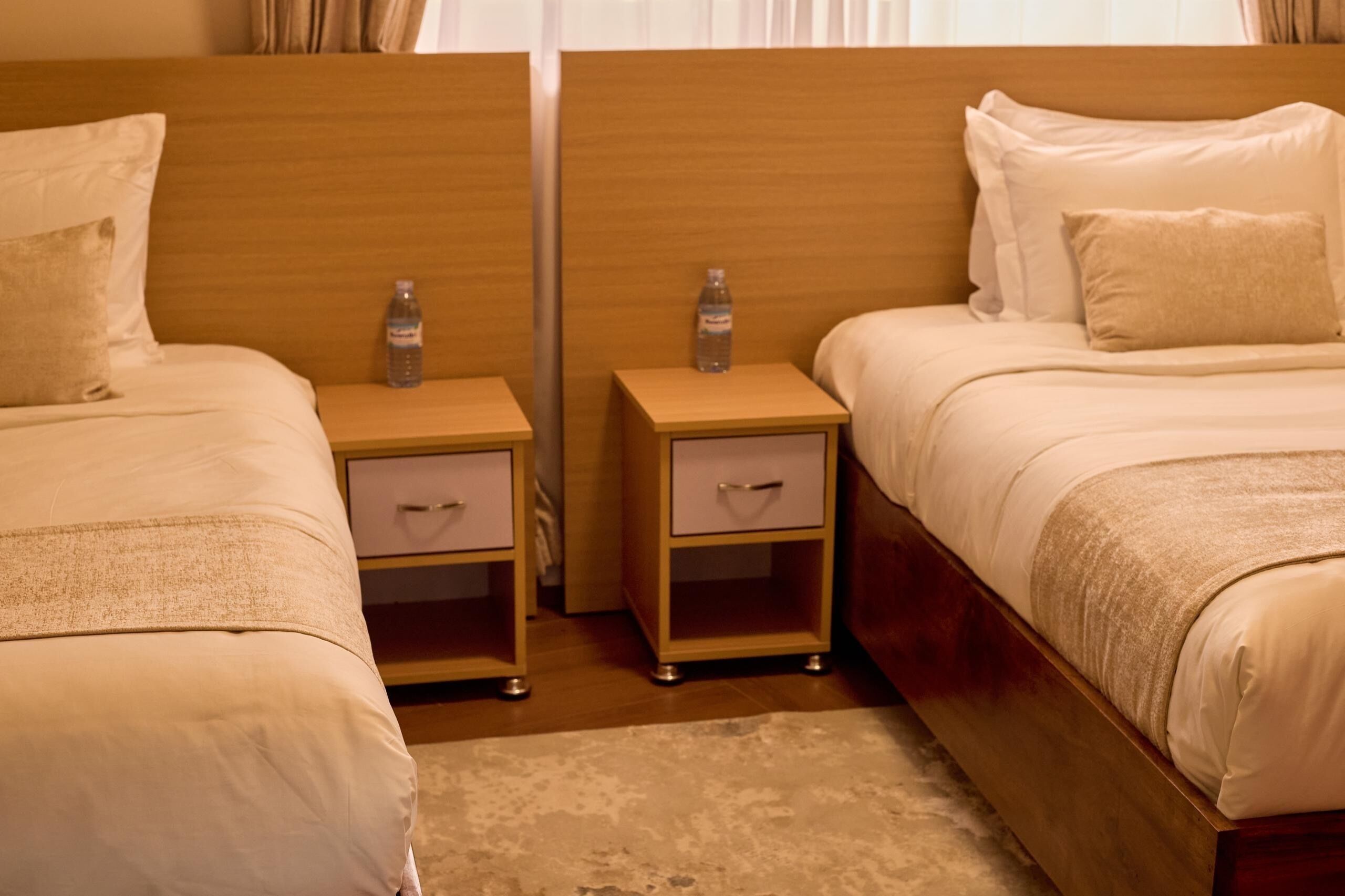 Een hotelkamer met twee eenpersoonsbedden, elk voorzien van een houten hoofdeinde, een nachtkastje met lade, en een fles water.