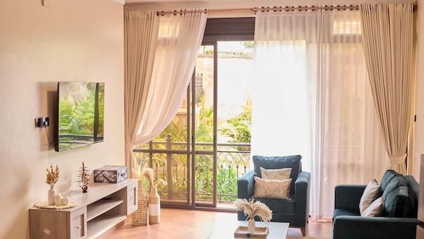 A living room with a TV mounted on the wall, a sofa, a coffee table, and a large window with curtains. - Sand Grains Residences 003 (Entebbe)