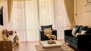 A living room with a dark green sofa, a black armchair, a white coffee table, and a wooden side table with decorative items. - Sand Grains Residences 003 (Entebbe)