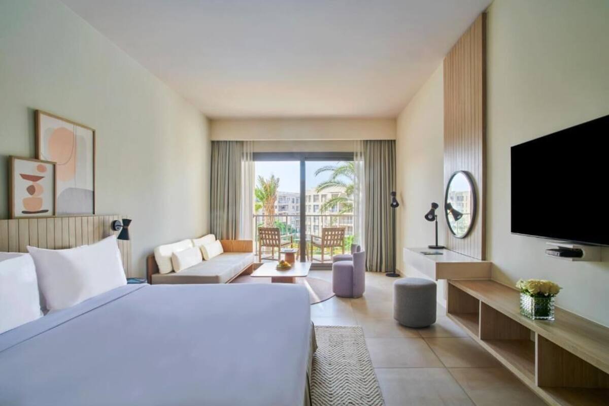 Une chambre d’hôtel équipée d’un lit, d’un canapé, d’une télévision et d’un balcon offrant une vue sur les palmiers.
