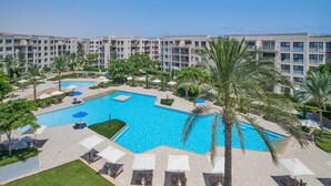 Une grande piscine extérieure entourée de palmiers et de chaises longues, avec un immeuble d’hôtel à plusieurs étages en arrière-plan.