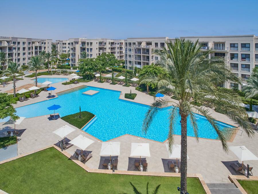 A large outdoor swimming pool surrounded by palm trees and lounge chairs, with a multi-story hotel building in the background.