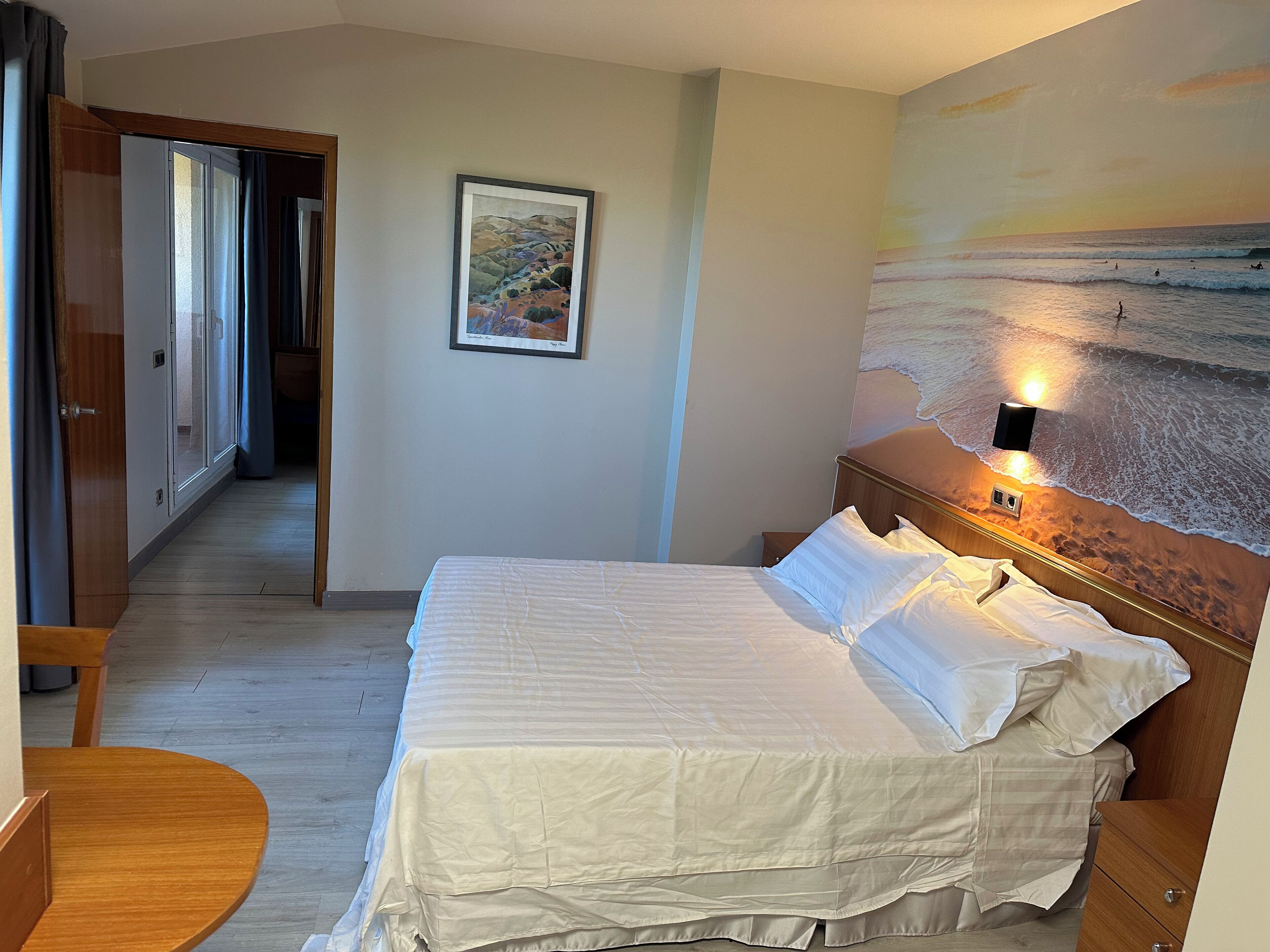Una camera d'albergo con un letto, un comodino con una lampada, un quadro appeso al muro e vista sulla spiaggia attraverso la finestra.