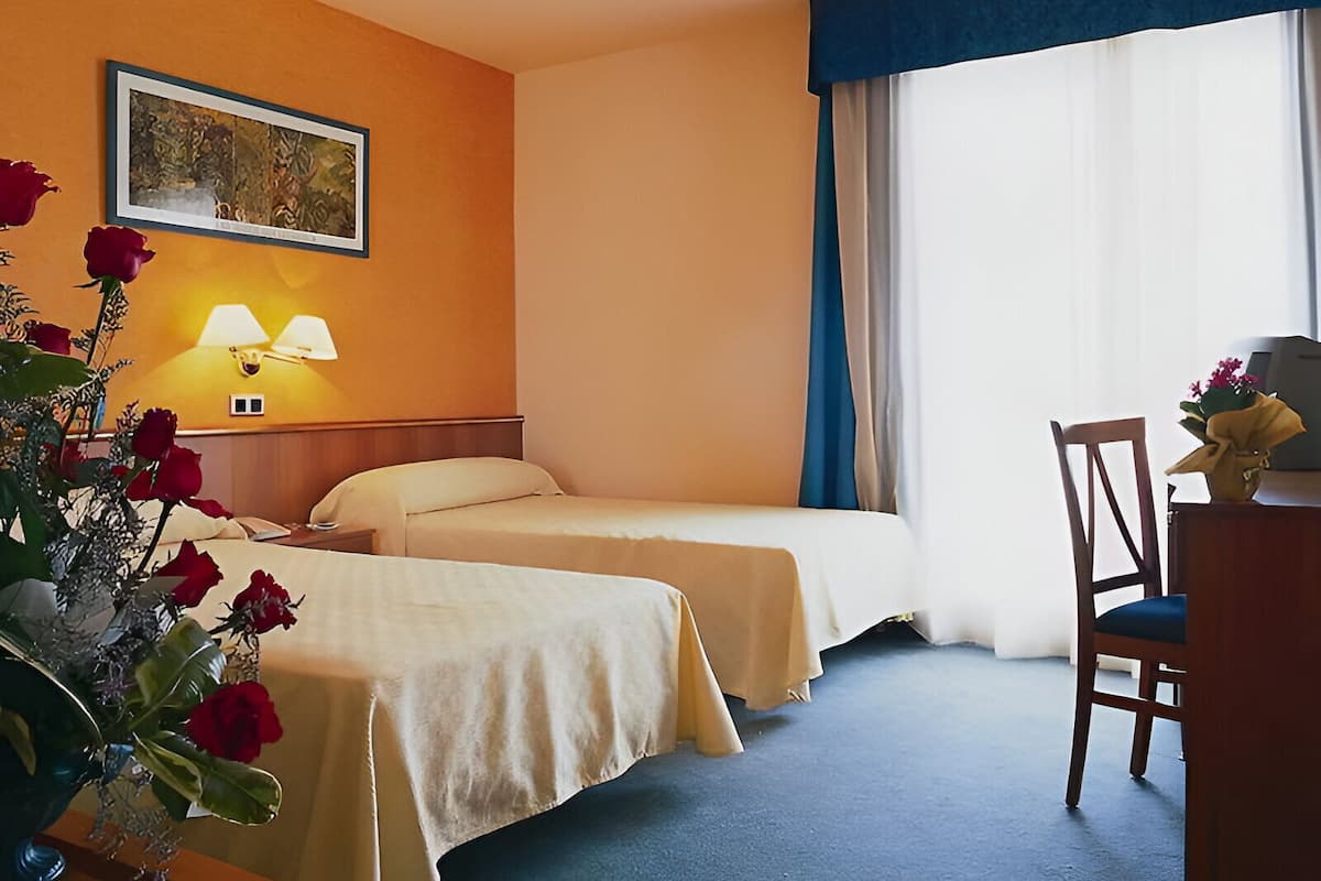 A hotel room with two beds, a desk, a chair, a lamp, and a painting on the wall.