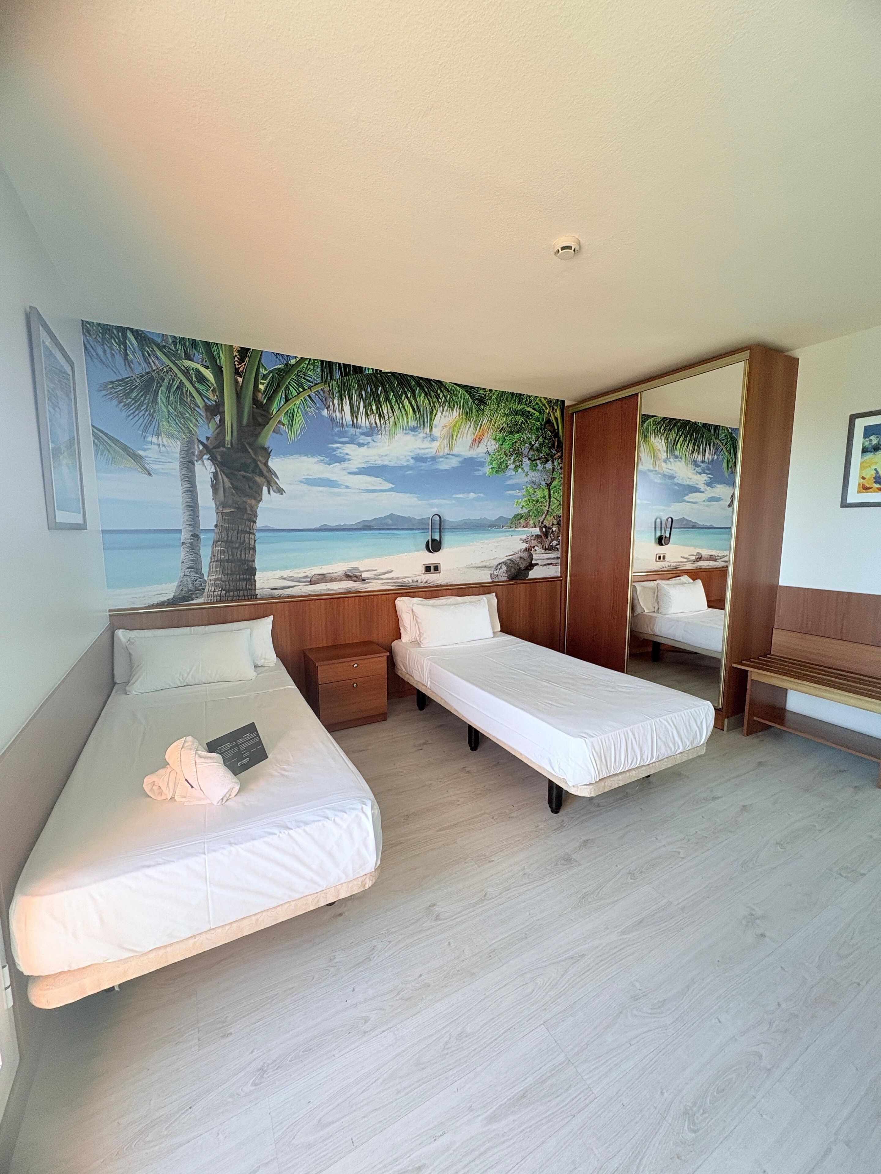 Habitación de hotel con dos camas individuales, una mesita de noche de madera, una pequeña mesa y un mural grande con motivos playeros en la pared.