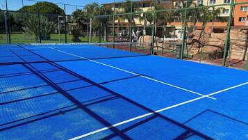 Een tennisbaan met een net, omgeven door een groene hekken en palmbomen.