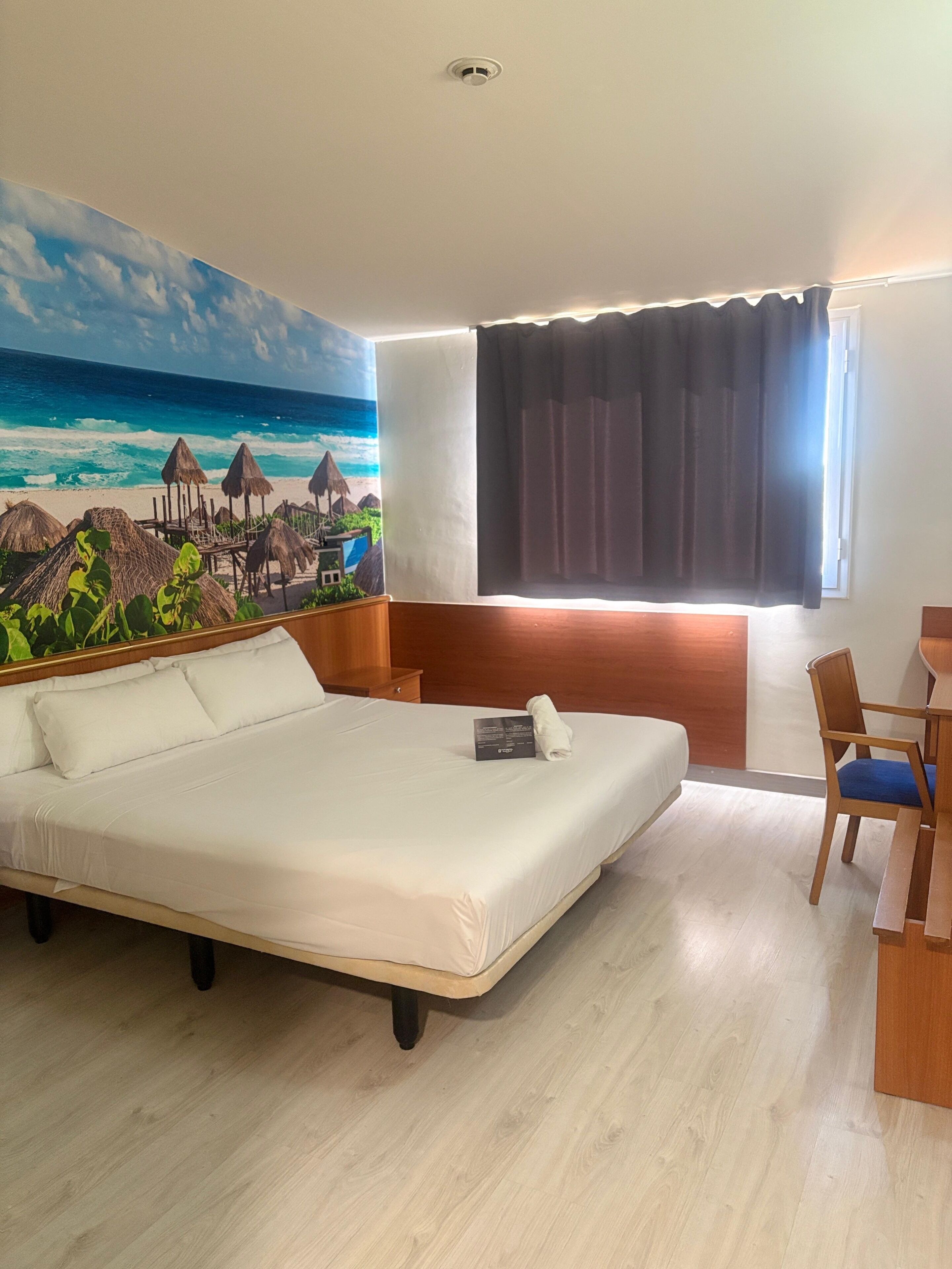 A hotel room with a large bed, a desk with a chair, a window with curtains, and a beach mural on the wall.