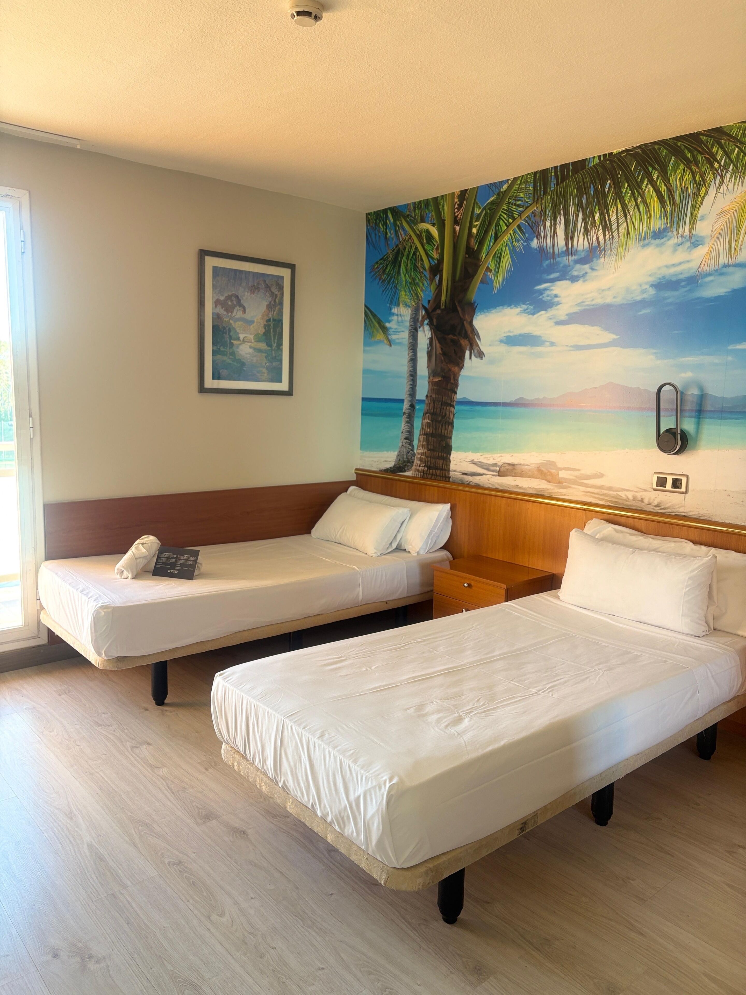 Habitación de hotel con dos camas individuales, cabecera de madera, un cuadro de una playa y una cámara de seguridad en el techo.