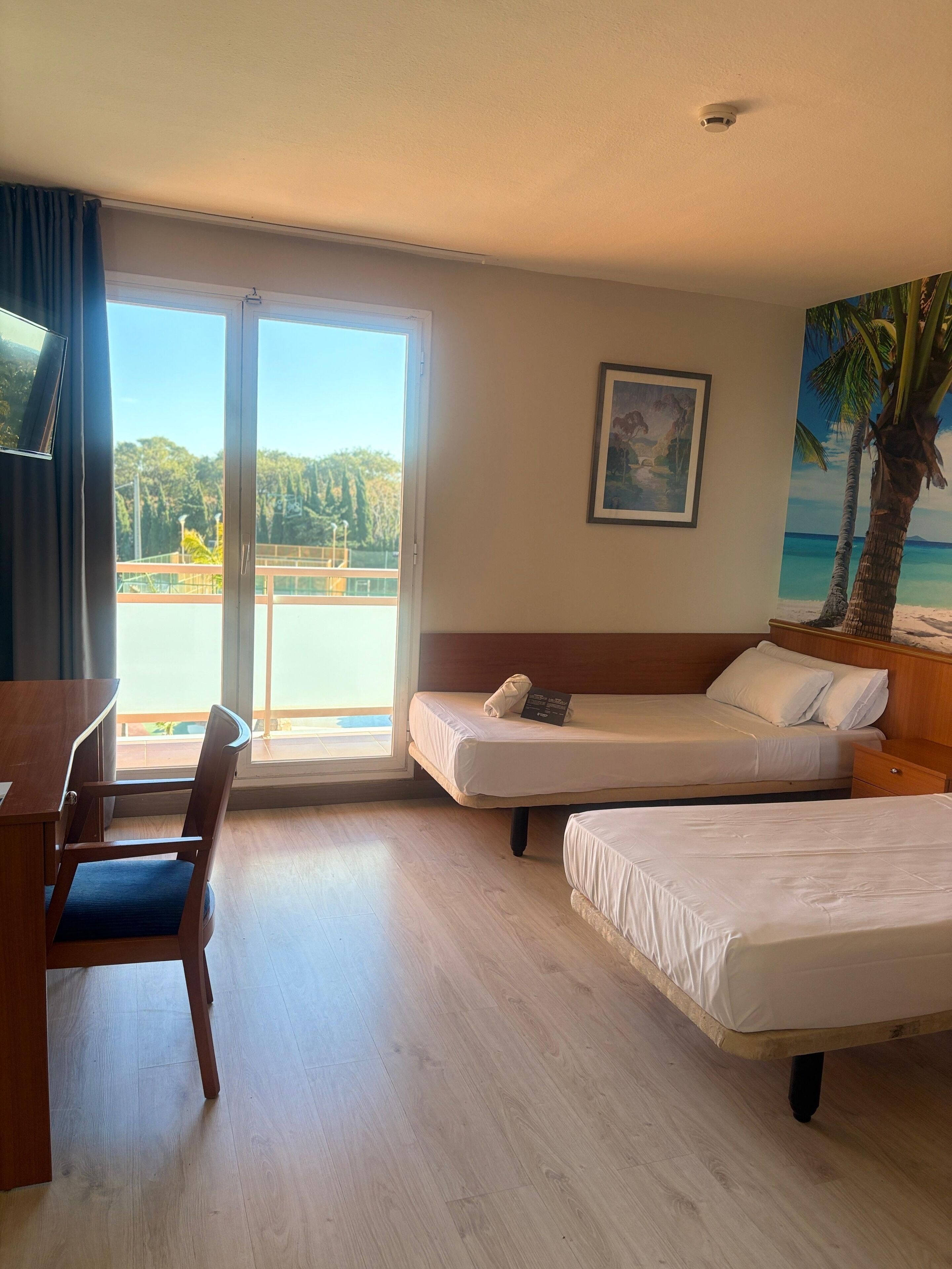 Habitación de hotel con dos camas, un escritorio y una silla. Amplia ventana con vista a una playa y palmeras.
