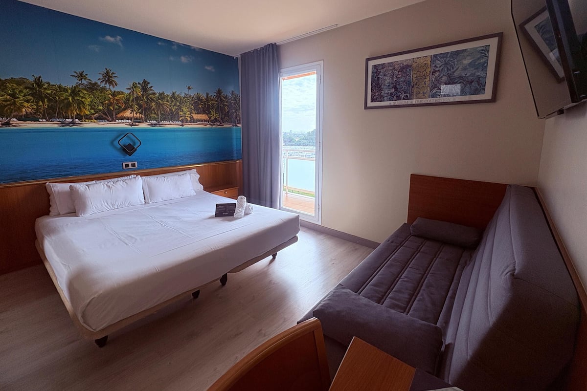 A hotel room with a large bed, a sofa, a painting, and a view of the sea through the window.