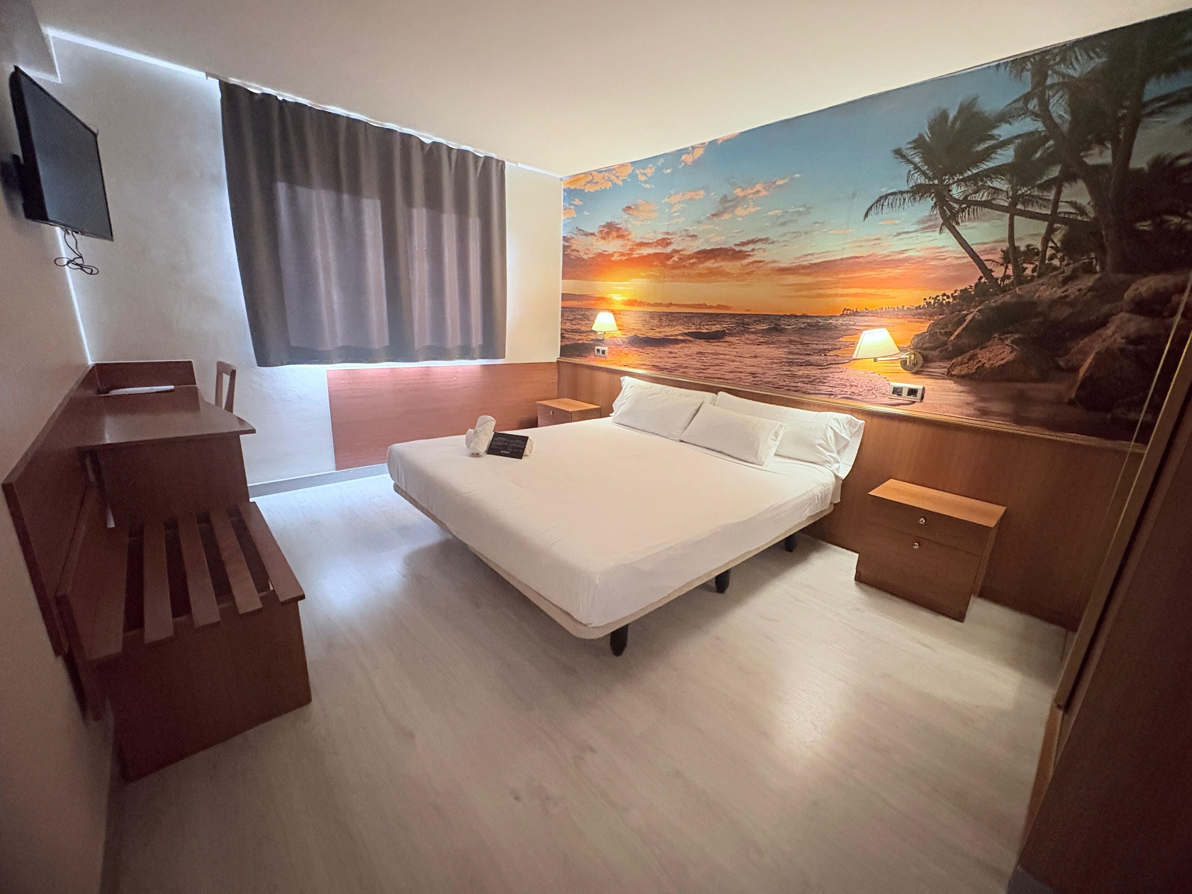 A hotel room with a bed, a desk, a TV, and a beach mural on the wall.