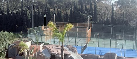 Zona de piscina con rocas y palmeras, rodeada por una valla y una cancha de tenis al fondo.