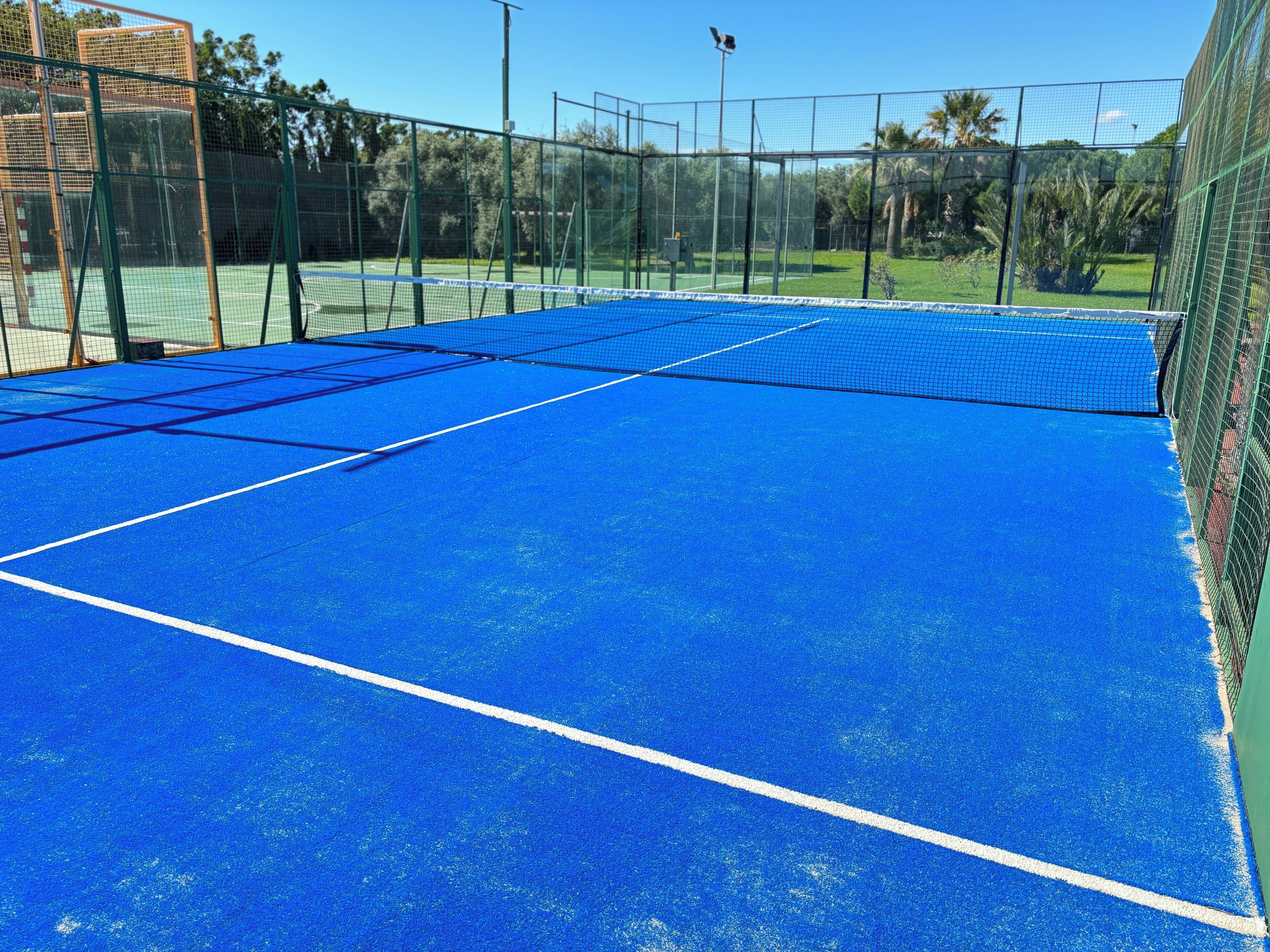 Uma quadra de tênis com superfície azul e linhas de marcação brancas, cercada por uma cerca verde.