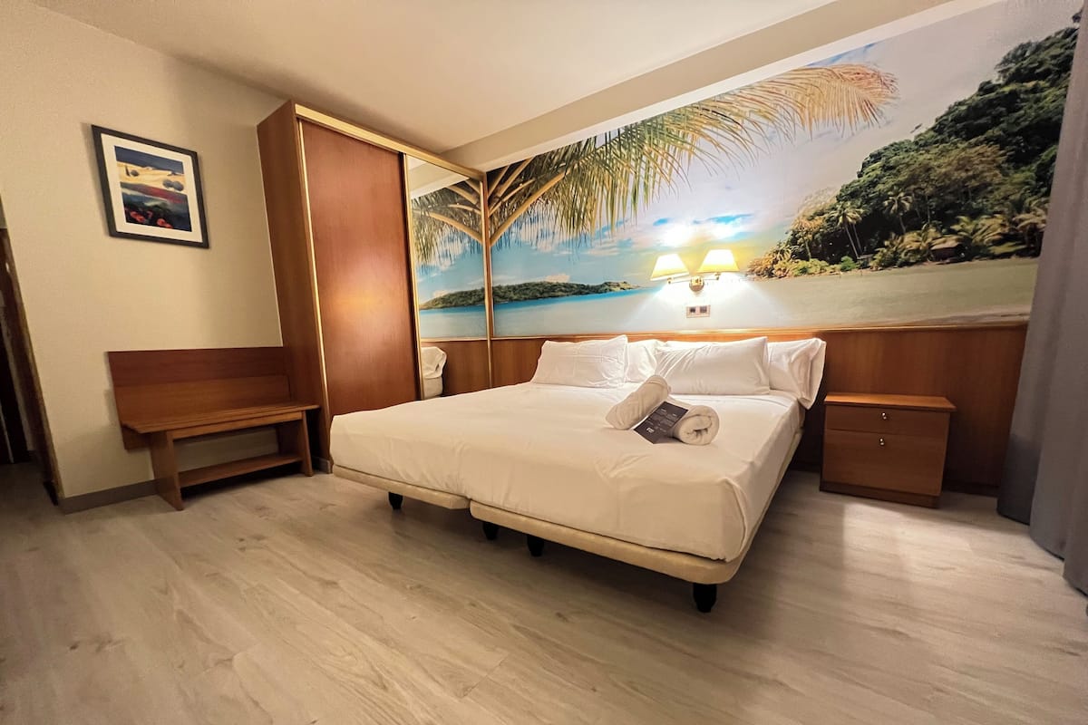 A hotel room with a bed, a bench, a nightstand, and a mural of a beach scene.