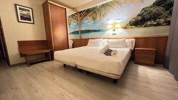 Habitación de hotel con cama, banca, mesita de noche y un mural de una playa.