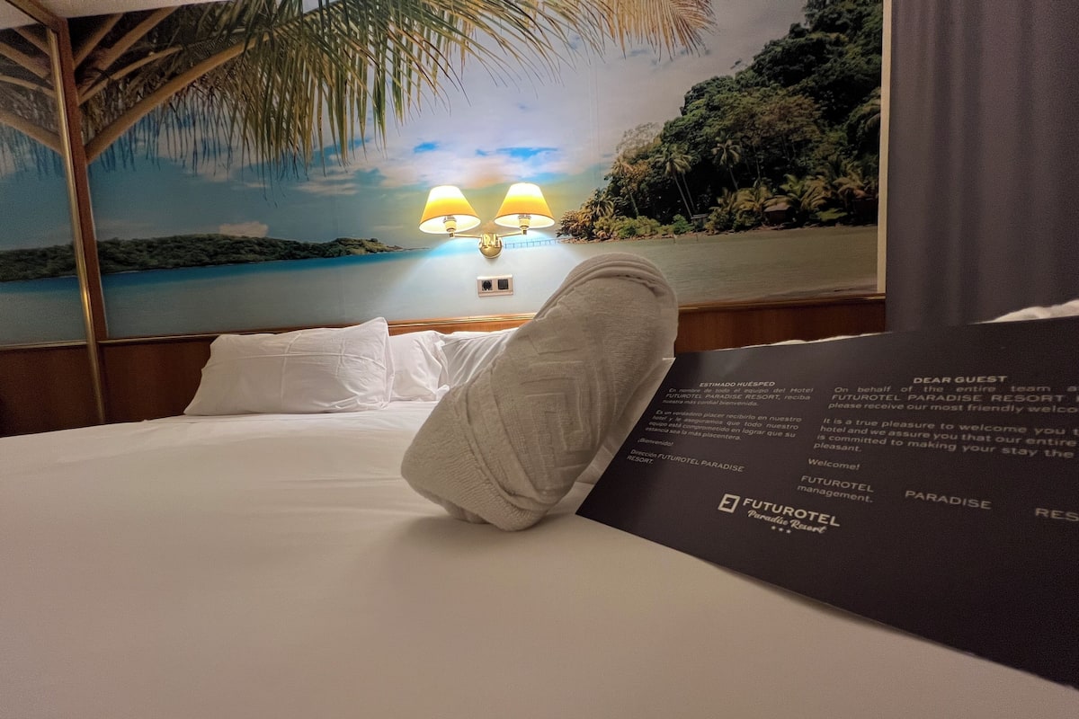 A hotel room with a bed, white pillows, and a wall mural of a beach scene. There are two wall-mounted lamps and a black sign with the hotel's name.