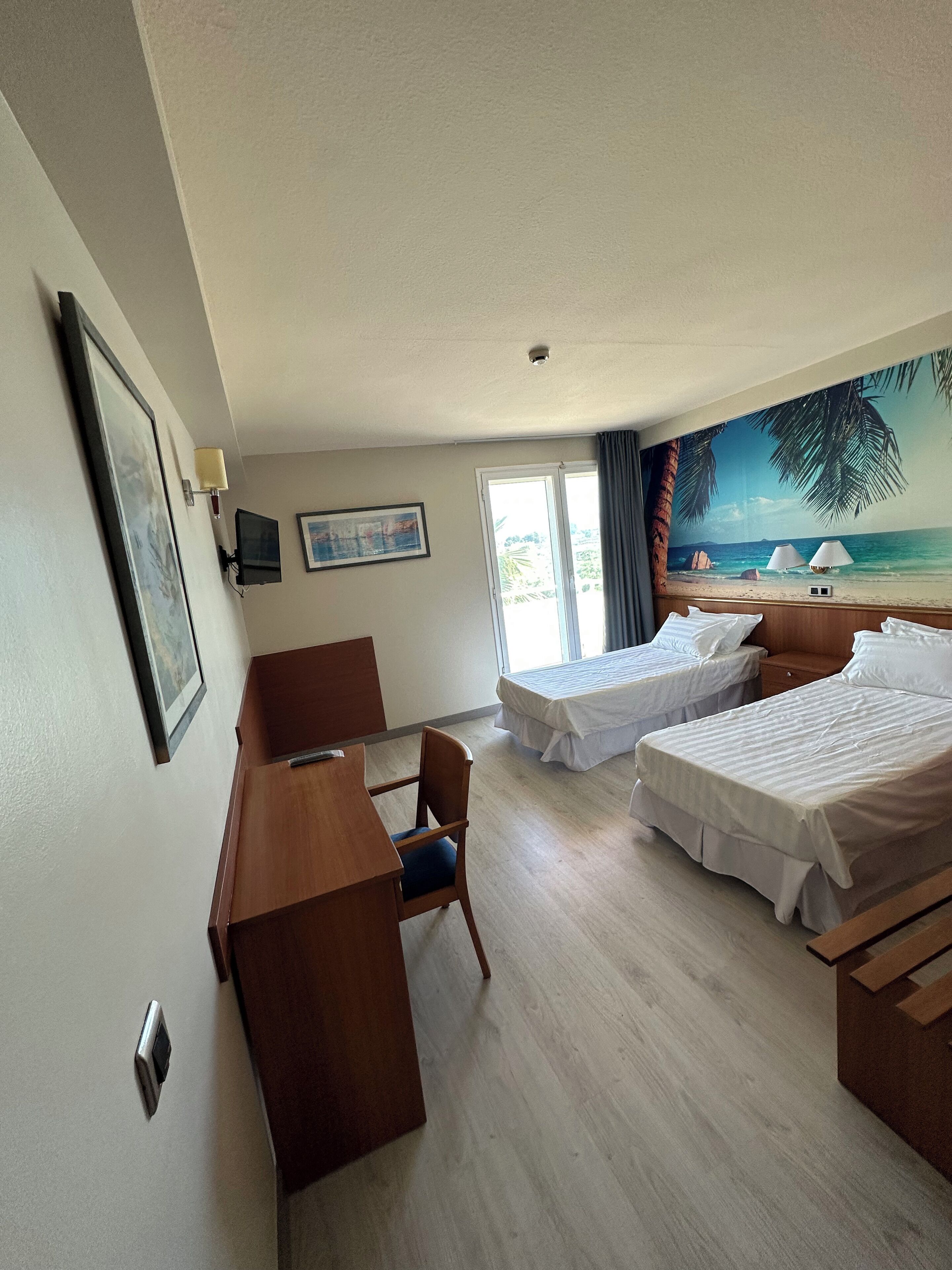 A hotel room with two beds, a desk, a chair, a TV, and a beach mural on the wall.