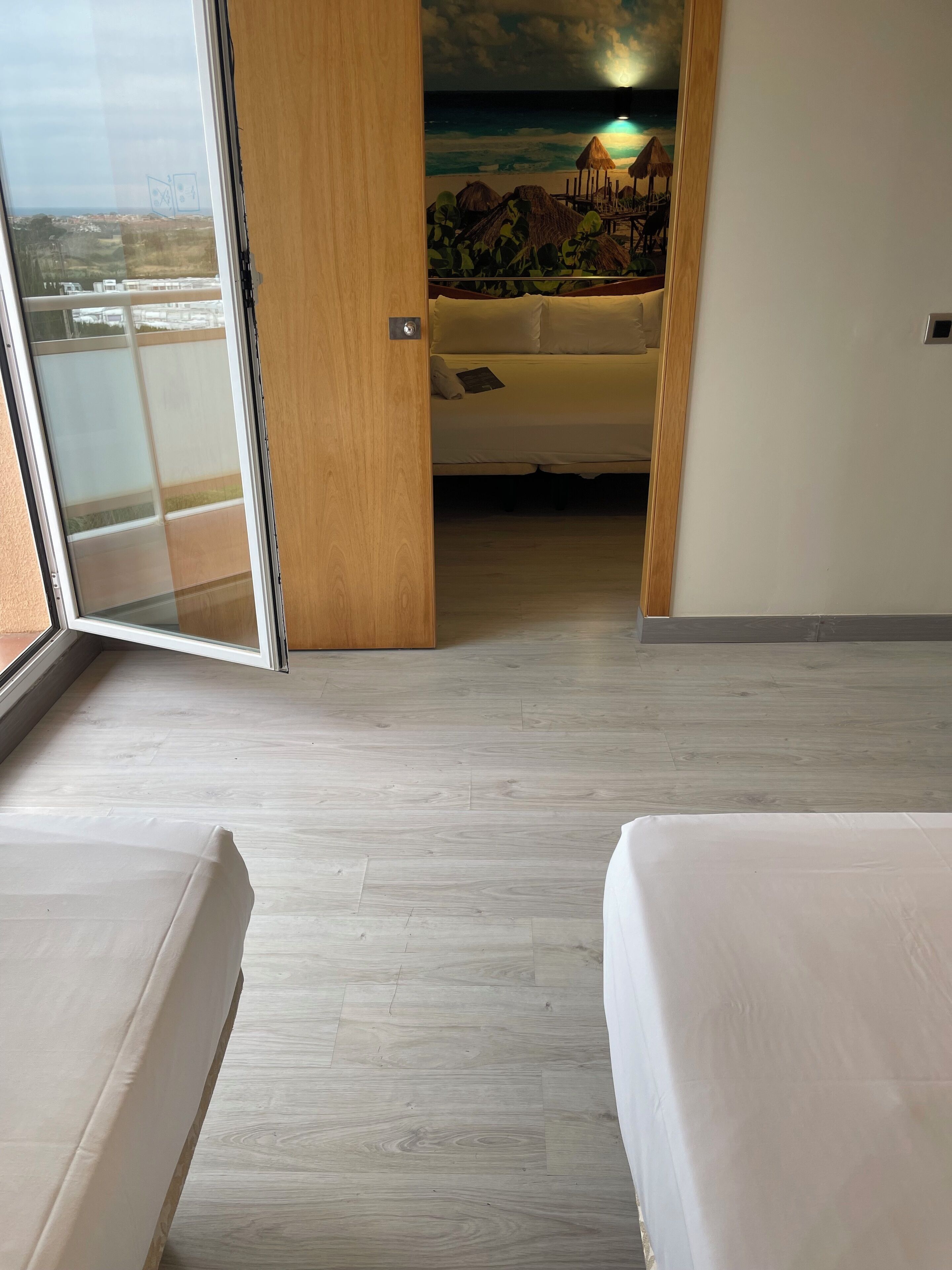 Una habitación de hotel con un amplio ventanal corredero que da a un balcón, una cama con colchón blanco y un mural con una playa en la pared.