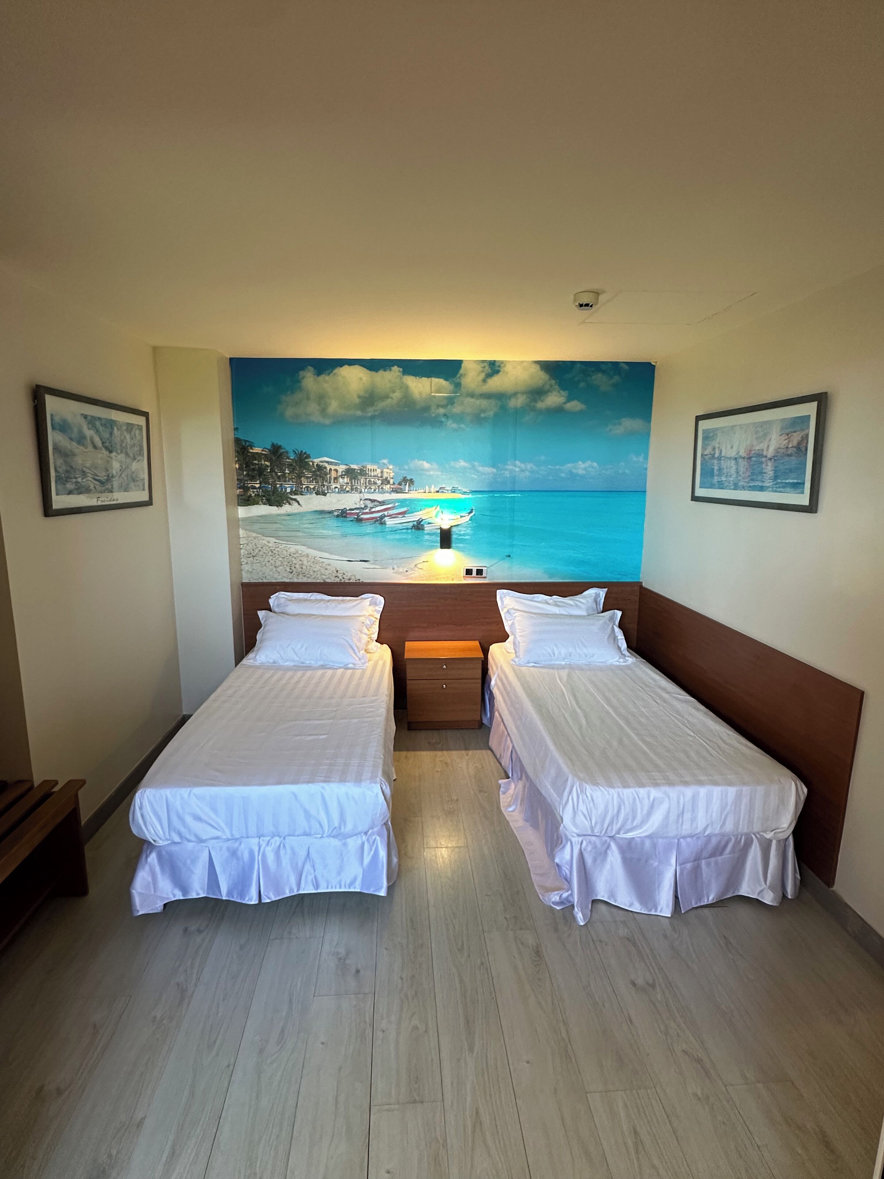 Ein Hotelzimmer mit zwei Einzelbetten, einem Holznachttisch und einem großen Wandbild mit Strandmotiv.