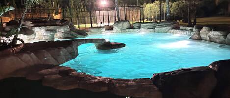 Una piscina nocturna con agua cristalina, rodeada de formaciones rocosas y palmeras.